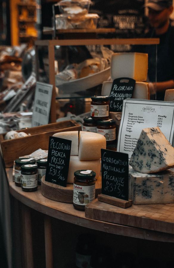 A specialty market counter of cut cheeses and jars with handwritten chalk price cards